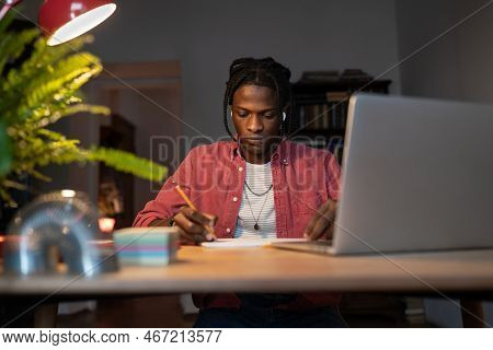 Focused Hardworking Young African American Guy Student Sitting At Desk Studying Under Lamp At Home T