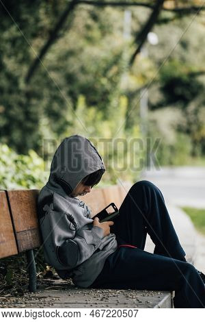 Boy With A Hood Sitting On A Bench Alone In A Park Looking The Smartphone