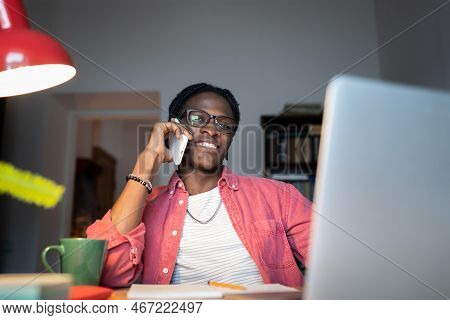 Friendly Smiling Young African Man Remote Worker Making Call Using Mobile Device While Working At Ho
