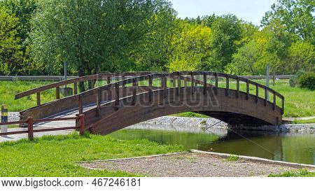 Wooden Arch Bridge Over Pond For Pedestrians In Park