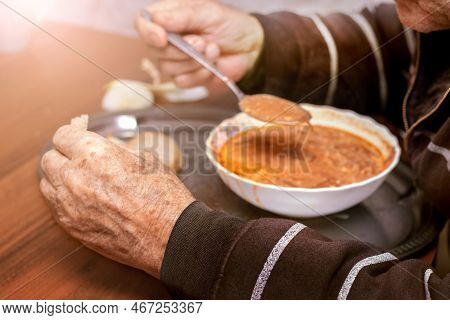 An Elderly Man Is Having Lunch, Eating Borscht. Grandfather Holds A Spoon And Bread Near A Plate Of 