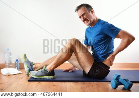 Man Doing Sports Indoors Complaining Of Lower Back Pain Sitting On A Mat. Side View