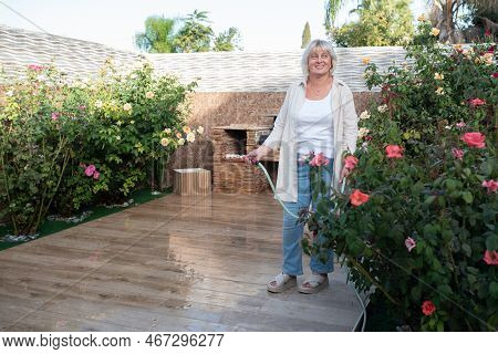 Portrait Of Smiling Senior Woman Holding Water Hose Watering Flowers At Backyard Garden. Beautiful H