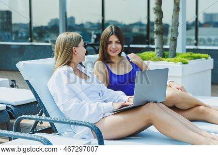 Two Beautiful Young Female Friends In Swimsuits Are Lying On Deckchairs By The Pool And Looking At I