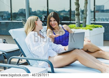 Two Beautiful Young Female Friends In Swimsuits Are Lying On Deckchairs By The Pool And Looking At I