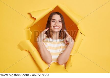 Indoor Shot Of Attractive Adorable Smiling Young Woman Wearing Striped T-shirt, Pointing At Her Whit