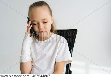 Portrait Of Sad Blonde Little Girl With Broken Arm Wrapped In Plaster Bandage Talking Smartphone, Sm