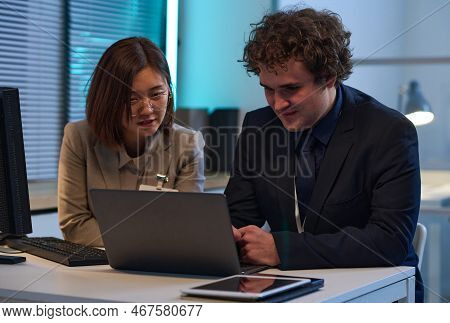 Young Colleagues Discussing Computer Program On Laptop While Working In Team In Office Till Late Eve