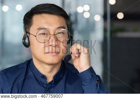 Close Up Photo. Portrait Of A Serious Asian Man In A Headset. He Looks Into The Camera, Holds The Mi