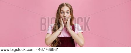 Cute Feminine Teenage Girl In Trendy Overalls With Tattoo On Arm Folding Lips In Wow, Touching Cheek