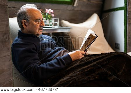 Elderly Man With A Mustache And Reading Glasses Covered With A Soft Warm Blanket Sit On A Bean Bag A