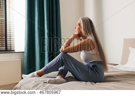 Senior Woman Sitting On Bed In Bedroom And Looking Away. Side View Of Pensive Mature Female.