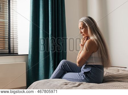 Sad Senior Woman Sitting On A Bed. Lonely Depressed Old Female Looking Down In The Bedroom.