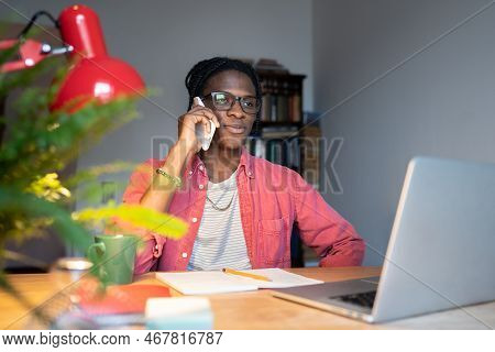 African Guy Wearing Glasses Talking On Phone While Sitting At Desk With Laptop, Distracted From Onli