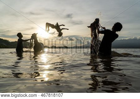 At Sunset, Three Silhouettes Of People Cleaning Racehorses On A Beach In Lariti Beach, Bima District
