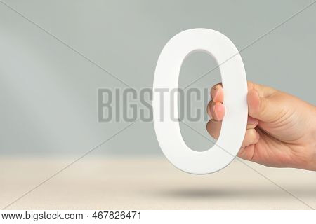 Numeral Zero In Hand. A Hand Holds A White Number Zero On A Blurred Background With Copy Space. Zero