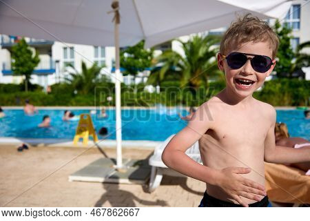 Cheerful Boy In Sunglasses Basks In The Sun Next To A Protective Umbrella After Swimming In The Pool