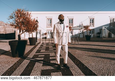 A Stylish Bald Black Man Dressed Elegantly In A White Suit, A Bow Tie, And Glasses, Is Standing On T