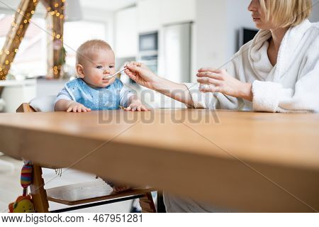 Mother Wearing Cosy Bathrope Spoon Feeding Her Baby Boy Child In Baby High Chair With Fruit Puree At
