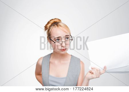 Young woman in glasses with papers in hand feels disappointed, grieved, sad and chagrined. Failure, bad luck, life or business problems. Grey background with copy space.