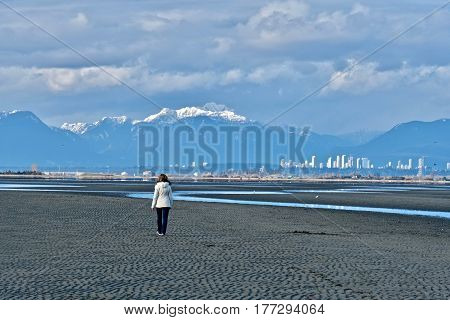 Woman walking on beach with downtown Vancouver and mountains view. Boundary Bay Regional Park in Tsawwassen. Great Vancouver. British Columbia, Canada.