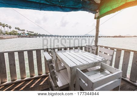 White dining table on corner of indoor beach terrace over water surface. Terrace surrounded by wooden wall and covered by colorful canvas.