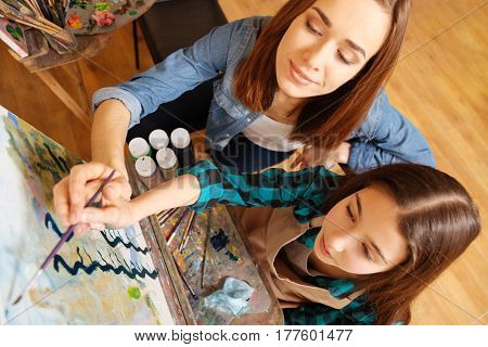 Conducting master class for kids. Cheerful charismatic young teacher sitting in the studio and having painting class while teaching children and expressing care and joy