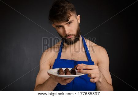 Hot sexy naked handsome man with chocolates chocolate candies. Smiling naked handsome man wearing dark blue apron holding a white plate with many sweets chocolate candies. Handsome confectioner.