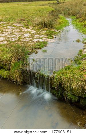 Small Stream Running On A Green Meadow With Stones And Smooth Water