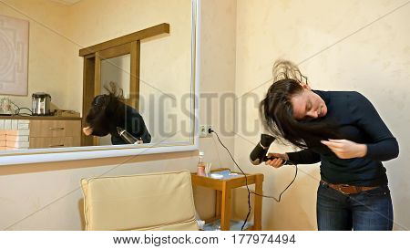Beautiful brunette woman drying long black silky hair with hairdryer and smartens up in front of mirror in make-up dressing room.