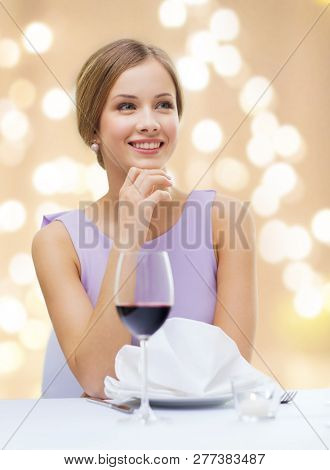 people and luxury concept - smiling young woman with glass of non-alcoholic wine waiting for date at restaurant over festive lights on beige background