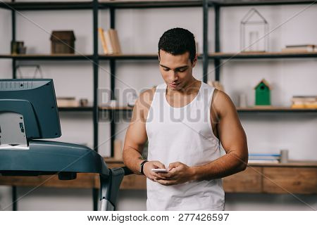 Handsome Strong Bi-racial Man Using Smartphone In Living Room Near Treadmill