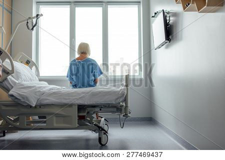 Back View Of Lonely Senior Woman Sitting On Bed In Hospital Ward With Copy Space