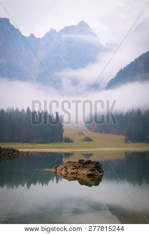 View Over Lake Laghi Di Fusine On A Foggy Morning With Some Rocks At The Beach And Mountain Range Ma