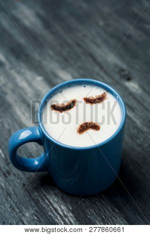 closeup of a blue cup of cappuccino with a sad face, drawn with cocoa powder on its milk foam, on a gay rustic wooden table with some blank space on top