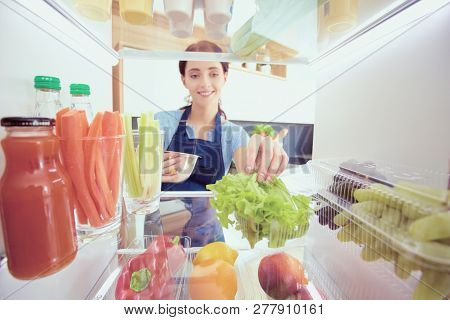 Portrait Of Female Standing Near Open Fridge Full Of Healthy Food, Vegetables And Fruits. Portrait O
