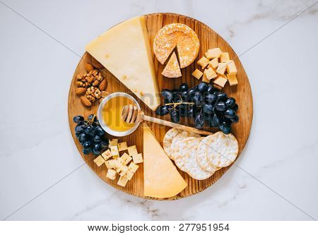 Flat Lay With Various Types Of Cheese, Grapes, Nuts, Honey And Cracker In Wooden Board On Marble