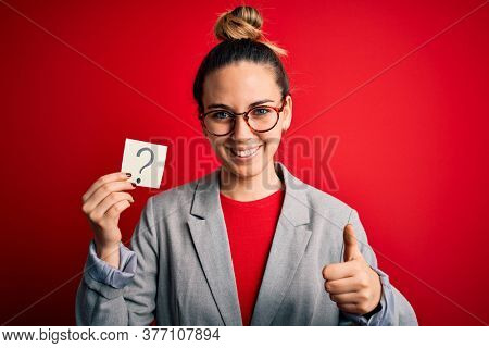Young beautiful blonde woman with blue eyes holding reminder paper with question mark happy with big smile doing ok sign, thumb up with fingers, excellent sign