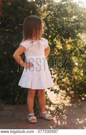 Beautiful Little Girl In White Dress Walking Outdoors On Back Yaer Of Home. Happy Kid