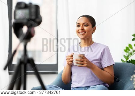 blogging, videoblog and people concept - happy smiling african american female video blogger with camera and cup of coffee videoblogging at home