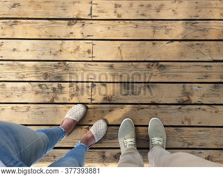 Two People Are Standing On A Wooden Surface. Top View Of Feet Shod In Light Summer Shoes.