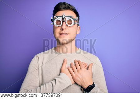 Young handsome caucasian man wearing optometrical glasses over purple background smiling with hands on chest with closed eyes and grateful gesture on face. Health concept.