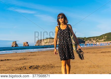A Young Brunette In A Black Dress And Sunglasses Walking Along The Beach In Hendaye, French Basque C