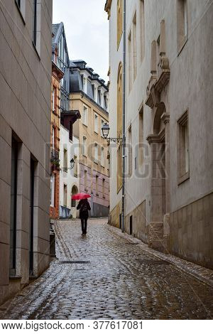 A Figure Walking Under An Umbrella On The Wet Cobblestones Among The Houses. Luxembourg City. Luxemb
