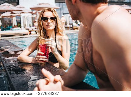Young Hot Couple Resting At Swimpool. View Of Sexy Gorgeous Woman Looking At Man And Drinking Cockta