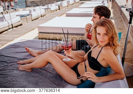 Young Hot Couple Resting At Swimpool. Woman And Man Lying Together And Resting Drinking Cold Drinks.
