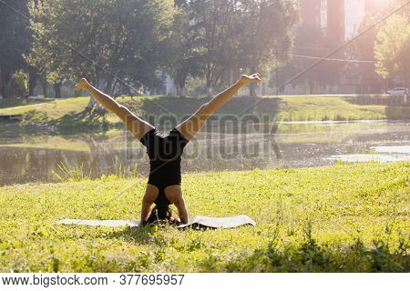 Young sporty attractive woman practicing yoga, doing headstand exercise, variation of upavishta konasana in sirsasana pose, working out, wearing black sportswear, in the park. Back view.