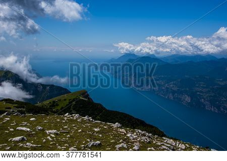 View Of Lake Garda Between Rocks, Clouds And Blue Sky On Monte Altissimo Di Nago In Trento, Italy