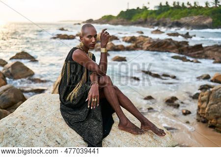 Androgynous Ethnic Fashion Model In Luxury Dress, Jewelry Sits On Rocks By Ocean. Gay Black Person I