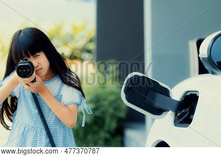 A Playful Girl Holding And Pointing An Ev Plug, A Home Charging Station Providing A Sustainable Powe